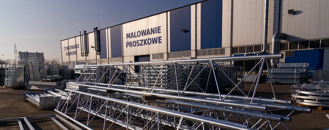 Widok na zakład cynkowania ogniowego Metaltech z napisem "Cynkowanie ogniowe" i "Malowanie proszkowe". Przed zakładem ocynkowane ogniowo konstrukcje stalowe